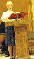 Person standing at a wooden lectern reading from a red book in a church-like setting