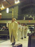 Parade of people in white robes carrying tall staffs through a large indoor hall