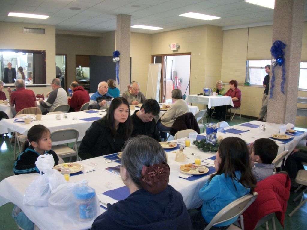 People seated at tables in a community hall, eating and talking during a meal gathering.