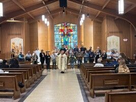 Church sanctuary with choir and clergy standing before a stained-glass window.