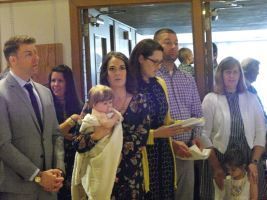People standing together indoors, including one holding a baby and another holding papers, in a doorway area