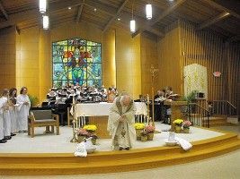 Church altar with clergy in white robes, flowers, and a stained-glass window behind the choir