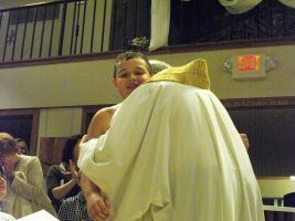 Person in a white robe and gold headpiece hugs another person in a hall.