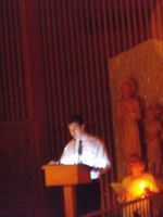 Person speaking at a podium on a dimly lit stage with red curtains and a statue in the background