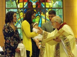 Baptism ceremony in a church, priest holding a baby as family members stand nearby.