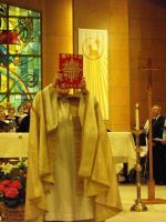 Clergy in white robes stands in a church aisle before a stained-glass window and altar.