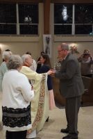 People in a hall exchanging a handshake during a ceremony, with an older man in a suit speaking to attendees.