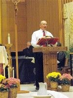 Man speaking at a church lectern during a service, with flowers and altar decorations nearby