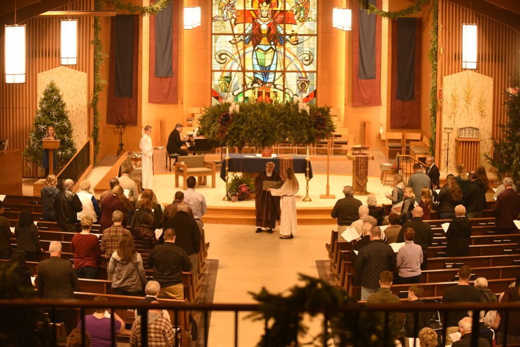 Church service with congregation seated in pews, altar flowers, and stained-glass window backdrop.