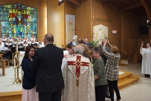 Church congregation gathered around clergy near the altar during a ceremony, with stained glass windows in the background