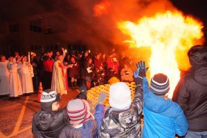 Crowd gathered at night around a large bonfire, with people in winter hats watching the flames