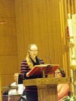 Speaker at a wooden podium reads from notes in a church or hall, with musicians seated behind.