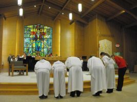 People in white robes bowing in a church with a stained-glass window and altar.