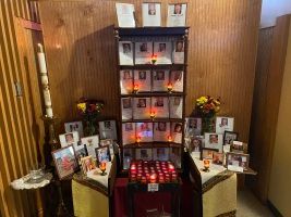 Memorial altar with framed photos, candles, flowers, and red lights in a wood-paneled room