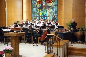 Choir and orchestra performing in a church sanctuary with stained glass and a pulpit.