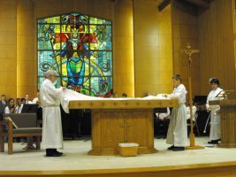 Clergy in white robes stand around an altar in a church with a colorful stained-glass window behind them