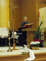 Person speaking at a church lectern with a red book, beside candles and floral arrangements.