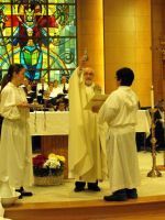 Priest and two attendants stand at the altar during a church service, with a stained-glass window behind them.