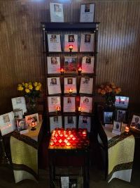 Memorial altar with framed photos, candles, flowers, and red votive lights against a wood-paneled wall