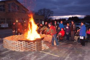 People gathered around a large bonfire in a brick-lined pit at dusk outdoors.