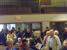 Crowd gathered in a hall, many holding papers, with a balcony and exit sign in the background