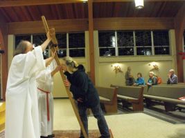Priest and person holding a wooden cross in a church aisle with seated congregation watching