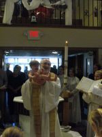Priest in white robes holding up a chalice during a church ceremony, with congregants standing behind.