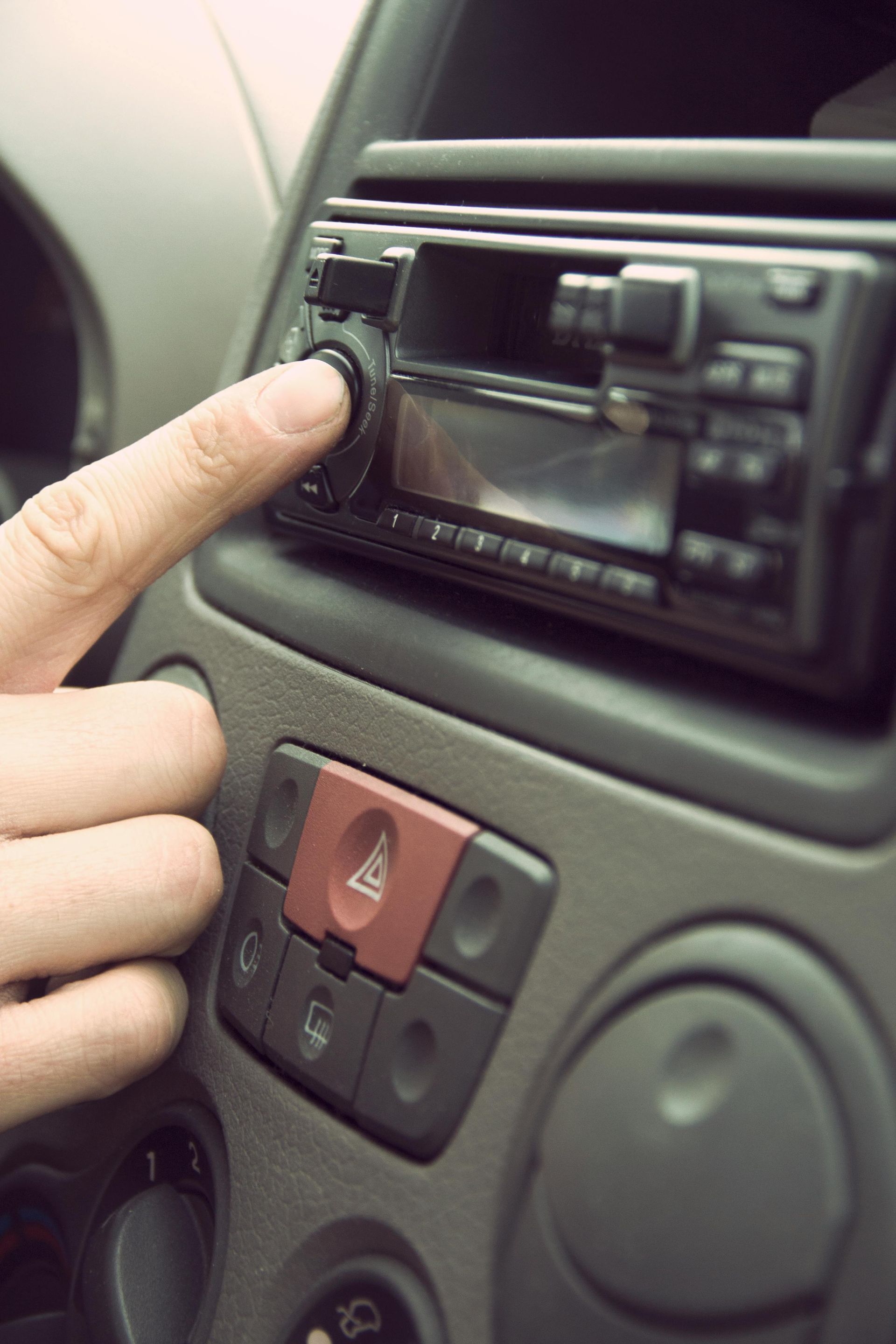 Hand turning dial on a car radio, next to hazard light button.