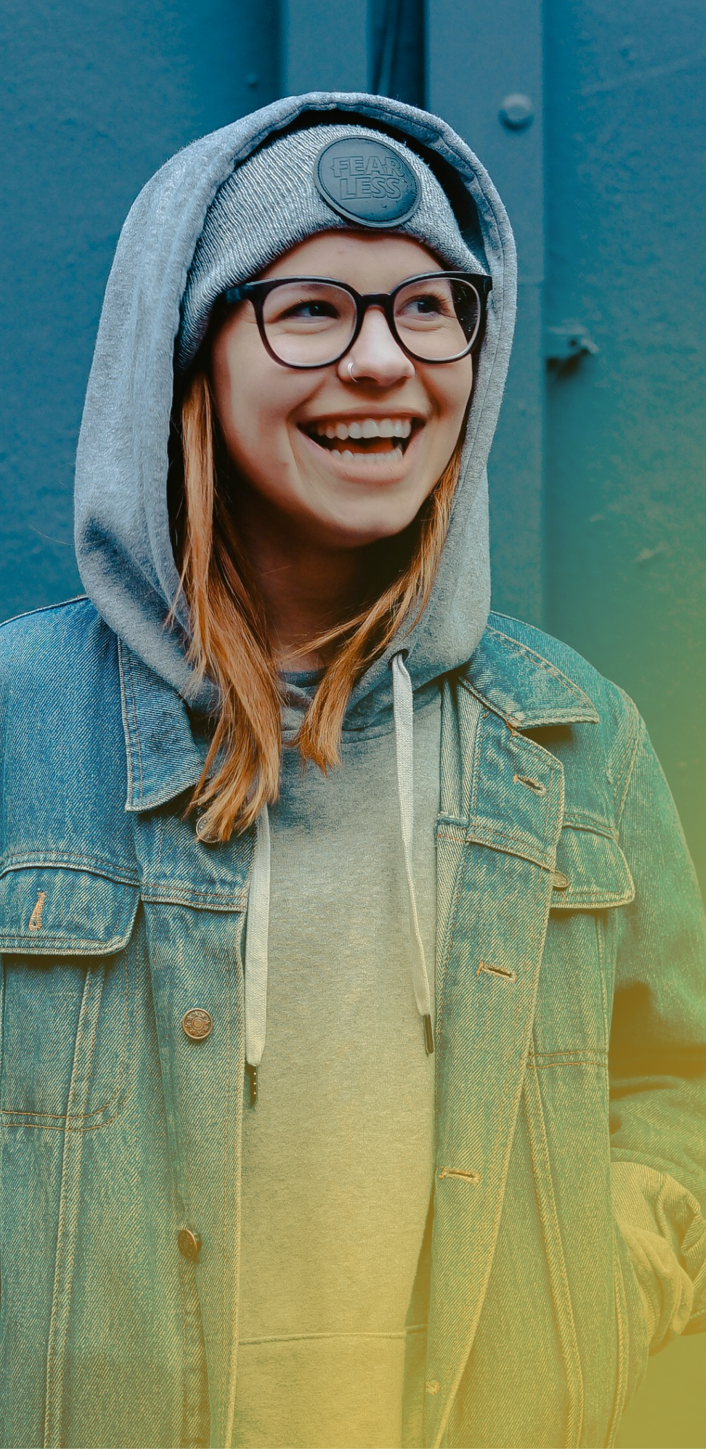 Smiling person in a gray knit hood and glasses against a blue wall, wearing a green coat