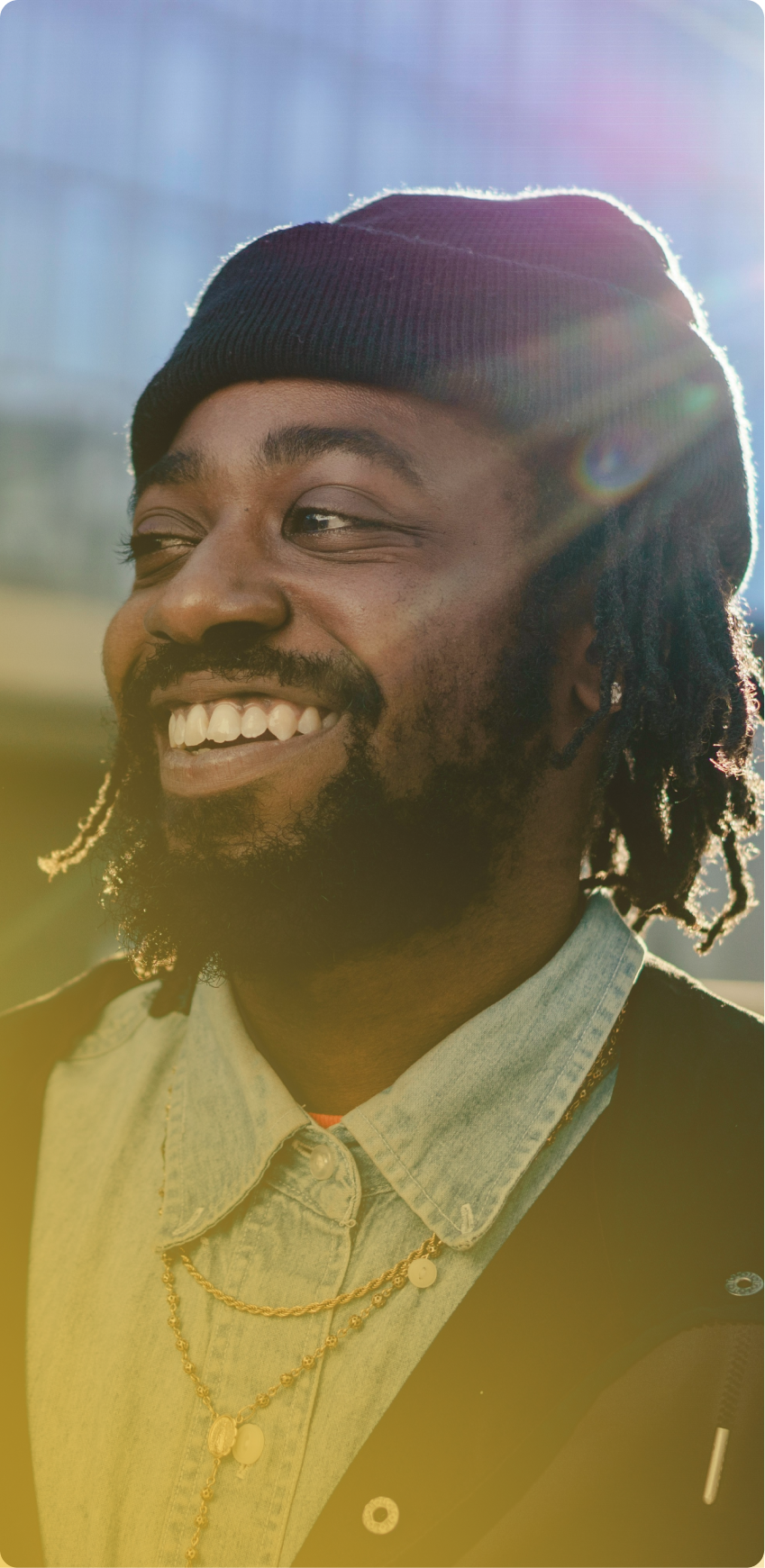 Smiling person in a beanie and layered shirts, standing outdoors in warm sunlight.