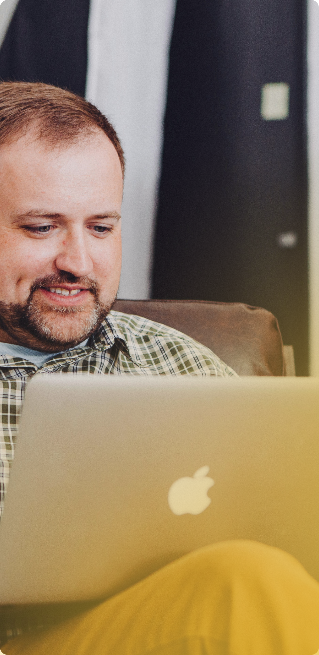 Man with a beard using a laptop on a couch, wearing a plaid shirt and mustard pants