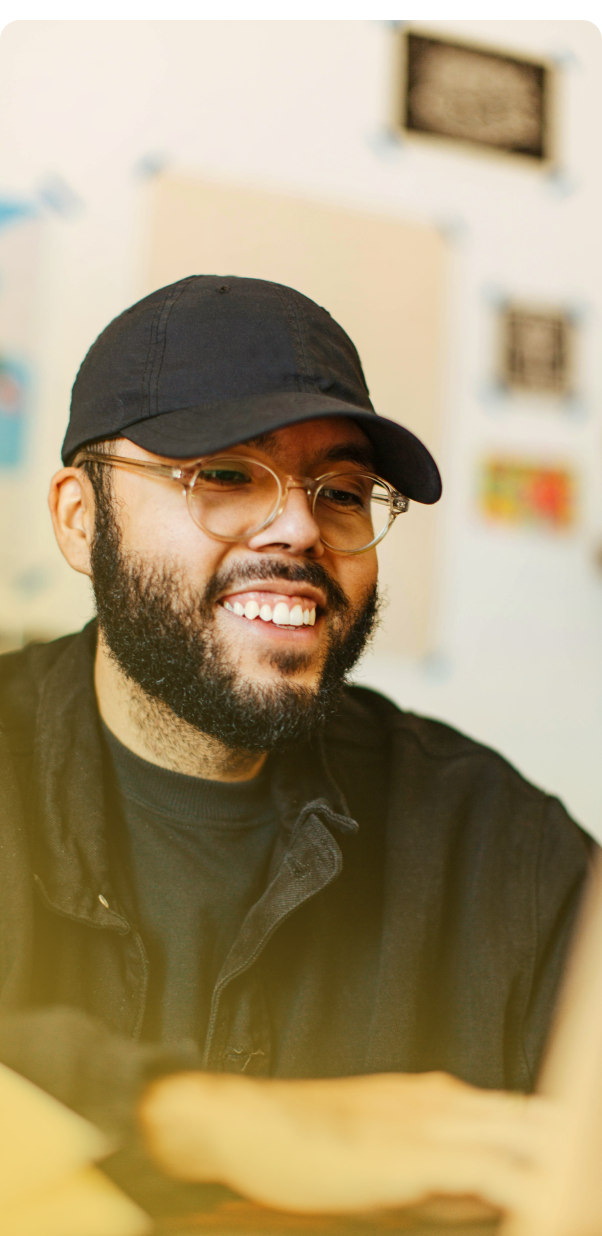 Smiling person in a black cap and glasses, sitting indoors with a warm blurred background