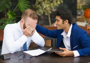 Two men in business attire at a table, one consoling the other who has his face in his hands.