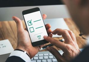 Hands holding a smartphone showing a checklist app with checked and unchecked boxes next to a keyboard.
