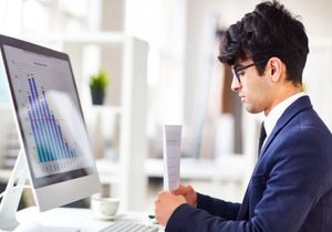 Man in a blue suit reviewing a document at a desk beside a desktop computer displaying charts