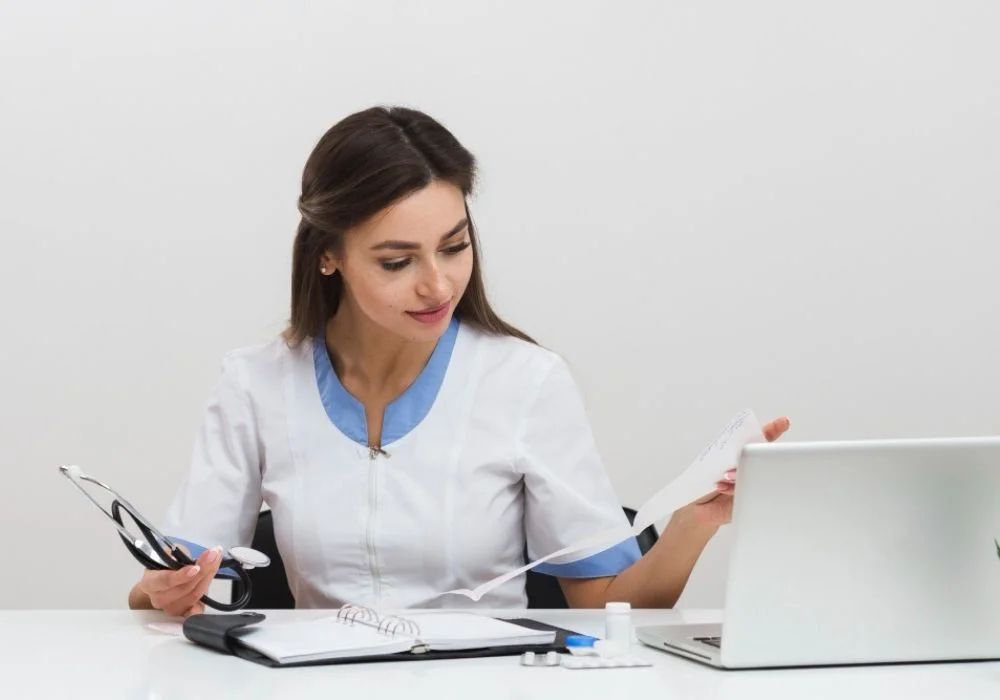 Woman in a white coat reviewing papers at a desk beside a laptop and office phone