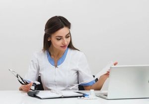 Woman in a white coat reviewing papers at a desk beside a laptop and office phone