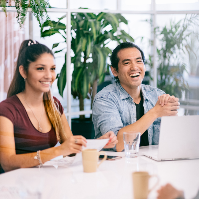 Two coworkers smiling at a table in a bright office, with laptops, coffee cups, and plants nearby