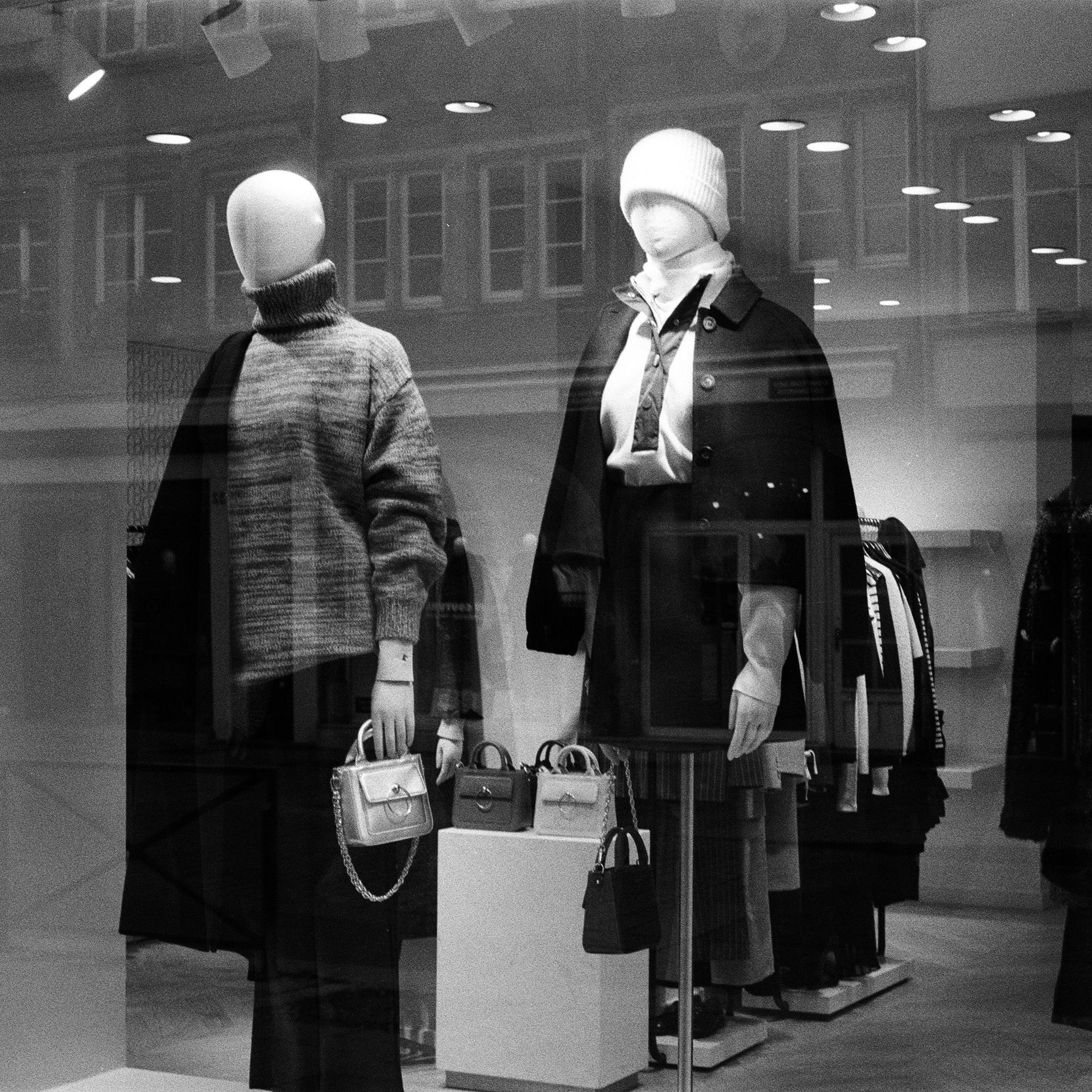 Store window display with mannequins wearing coats and handbags, reflected in glass.
