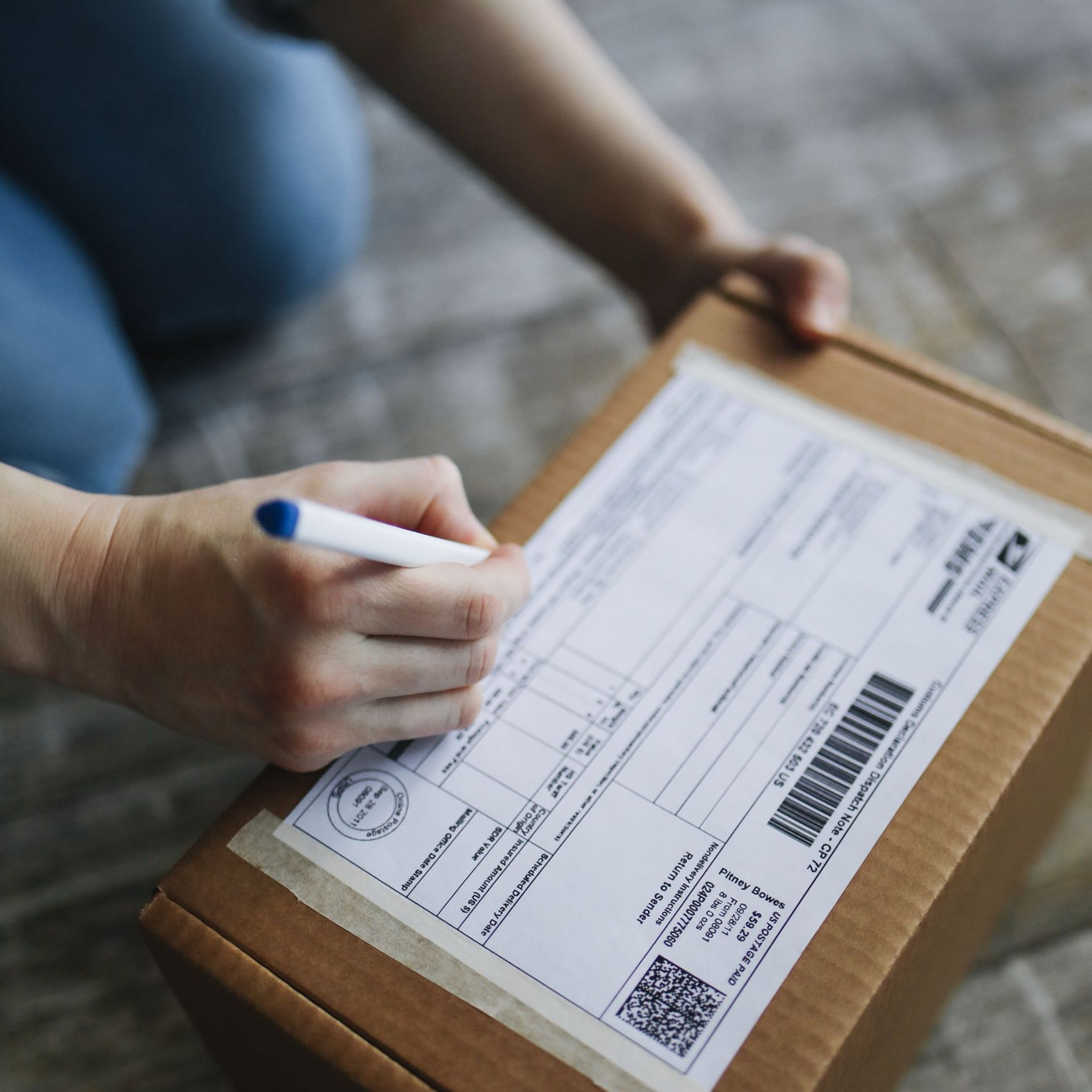 Person writing on a shipping label attached to a cardboard parcel on the floor.