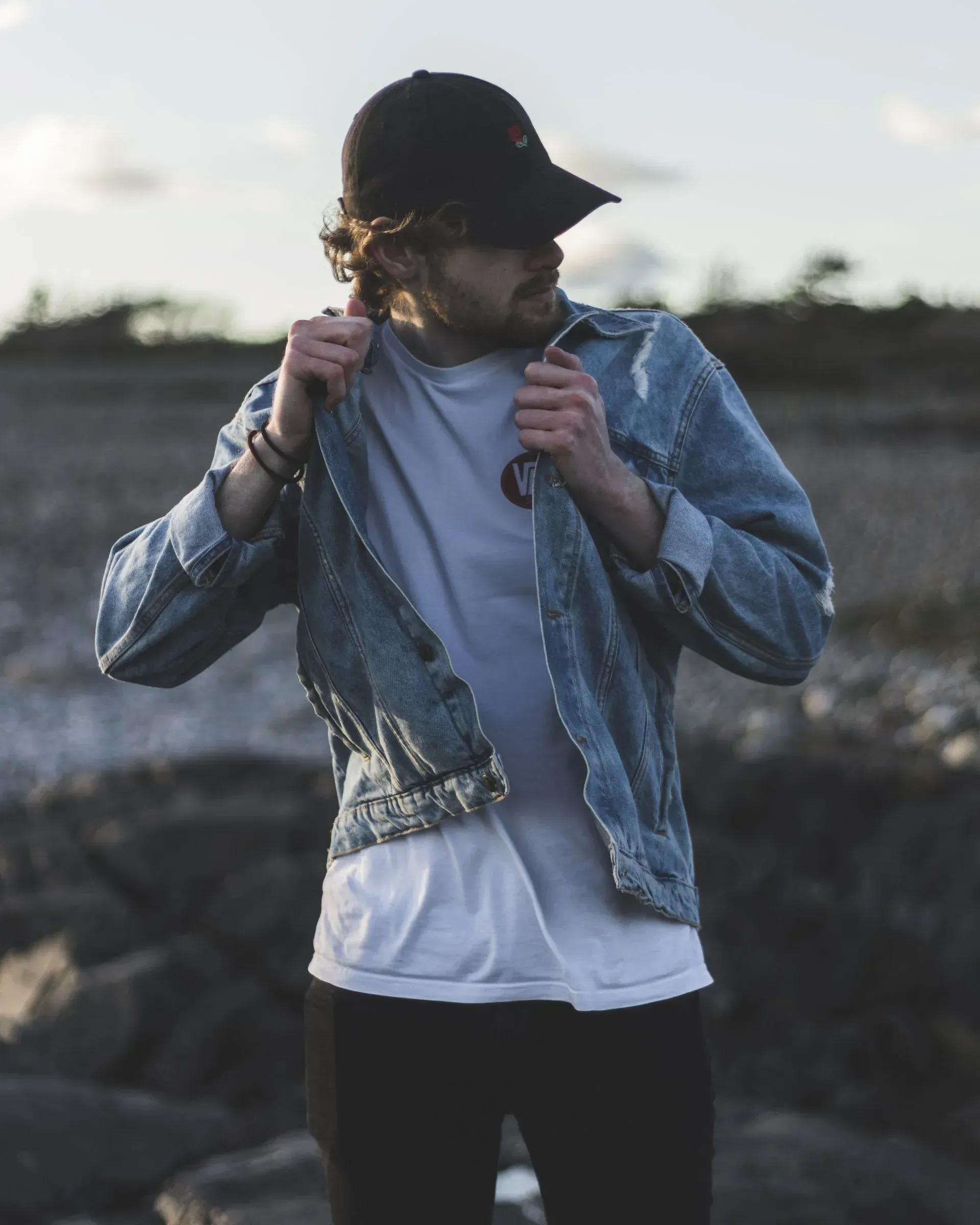 un uomo che indossa una giacca di jeans e un cappello è in piedi su una spiaggia rocciosa .