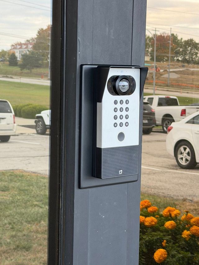 Apartment intercom with keypad mounted on a dark pillar near a parking lot and orange flowers