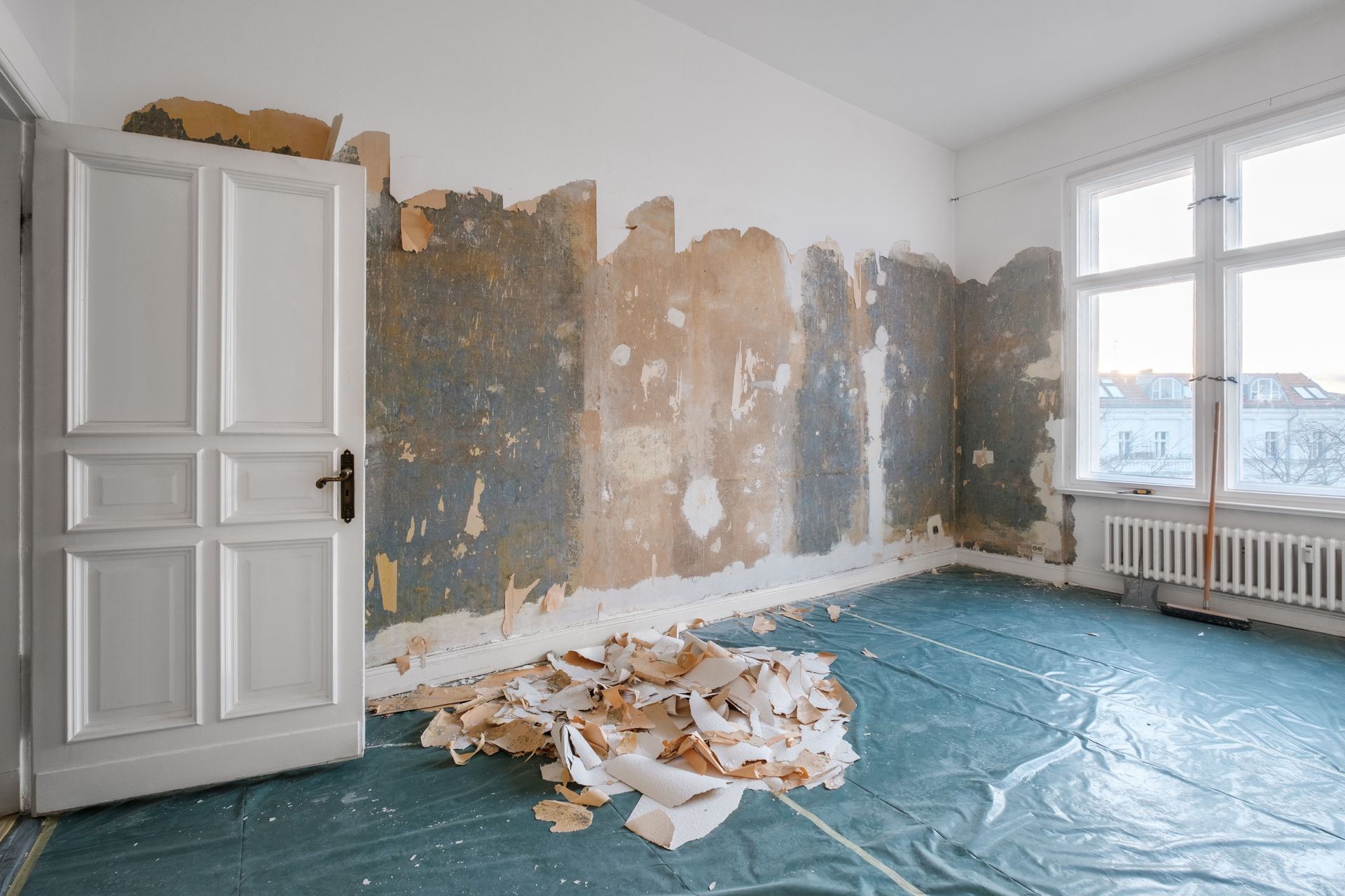 Renovierungsraum mit abblätternden Wänden, Boden bedeckt mit Schutzfolie und Bauschutt, und einem weißen Schrank