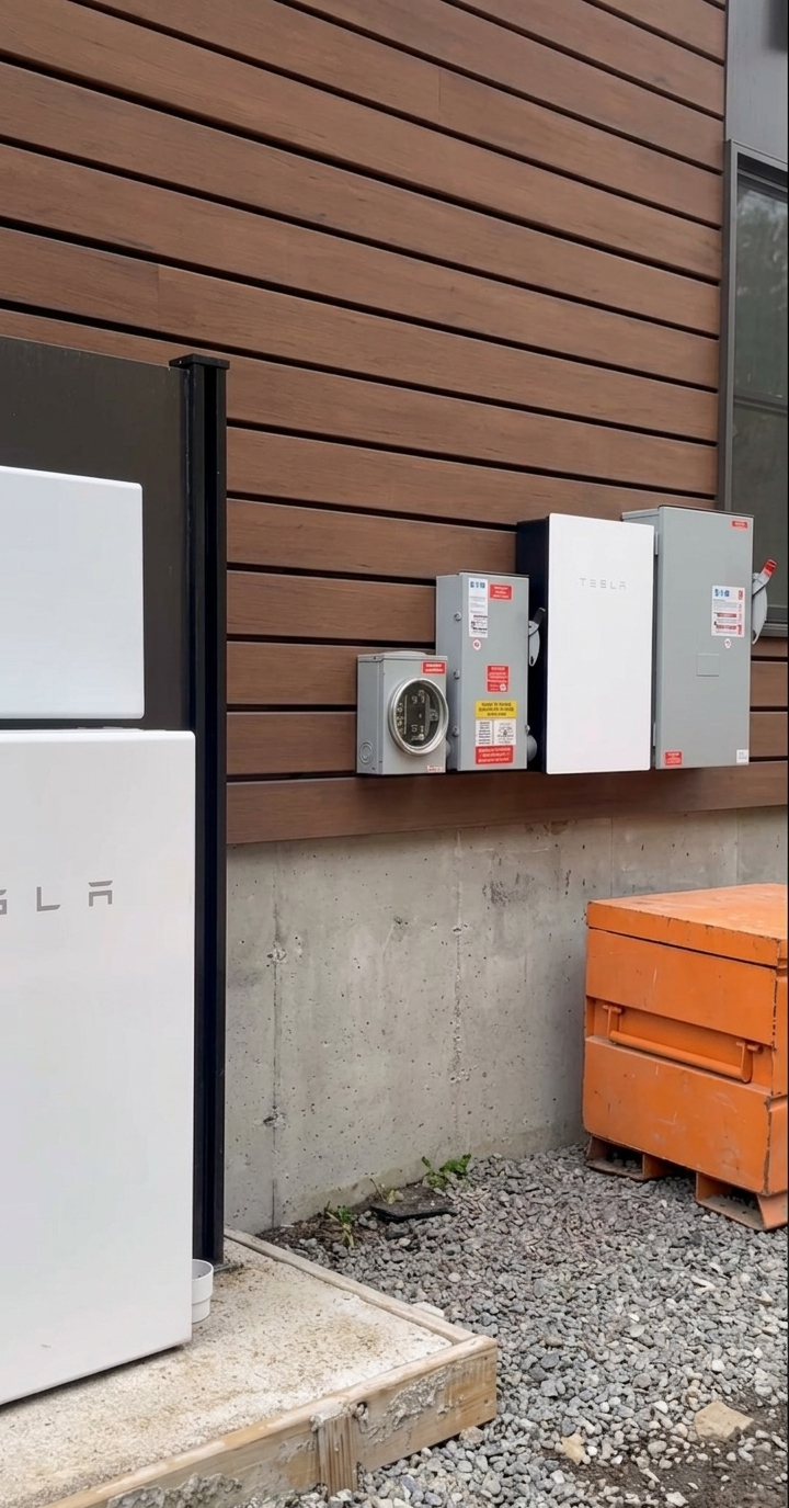 A Tesla Powerwall and electrical equipment mounted on a wooden building exterior above a gravel ground surface.
