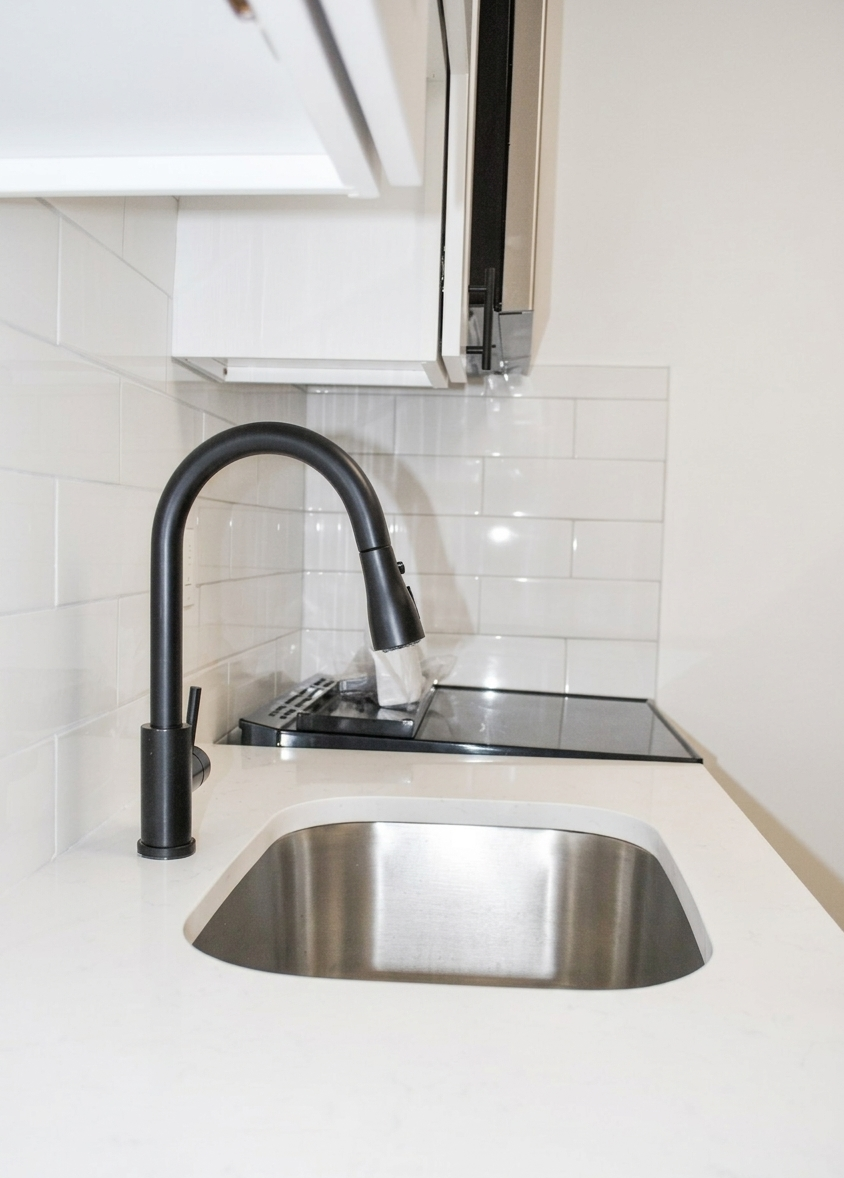 A close-up view of a matte black kitchen faucet and stainless steel sink set against white cabinets and a subway tile wall.