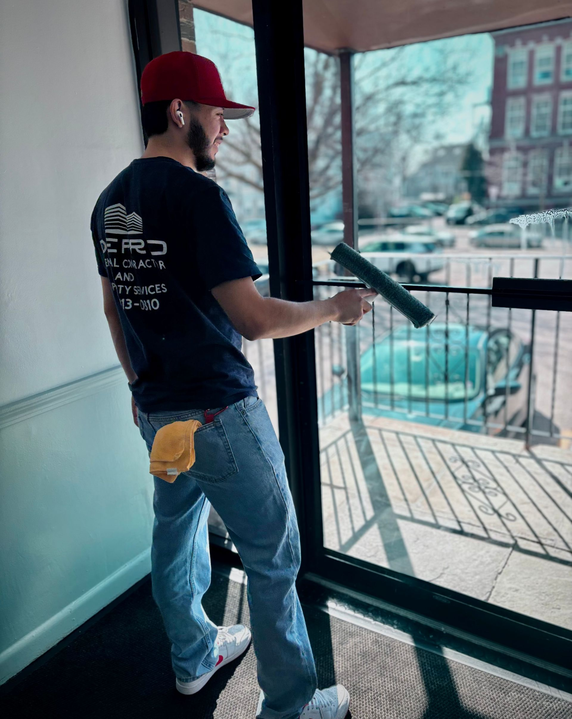 A professional window cleaner wearing a red cap and dark t-shirt uses a squeegee to clean a glass door.