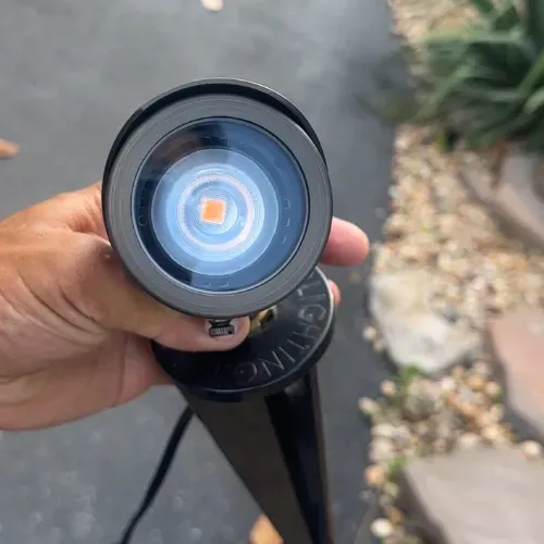 A hand holding a brass LED landscape light with a bright orange center against an outdoor setting.