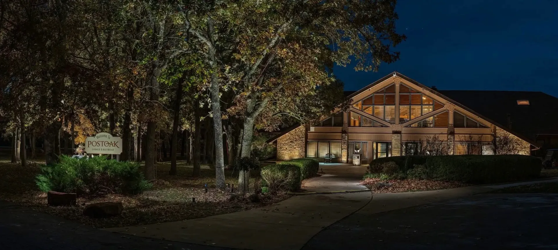 Nighttime exterior of a wood-beamed building with trees, lights and a sign illuminated.