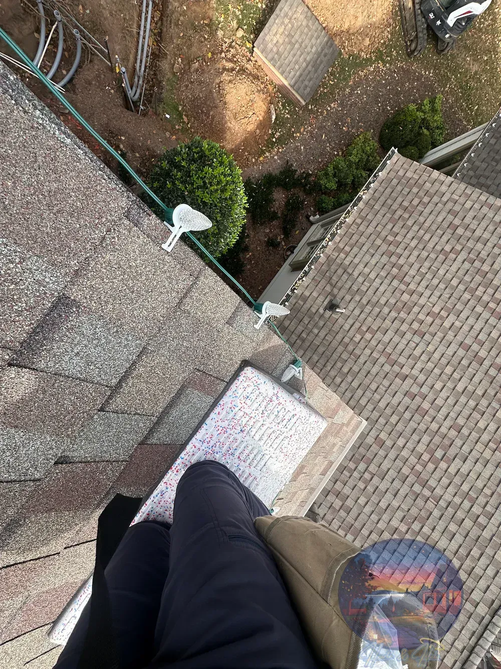 A first-person view looking down from a residential shingled roof towards the ground, showing legs and a tool bag.