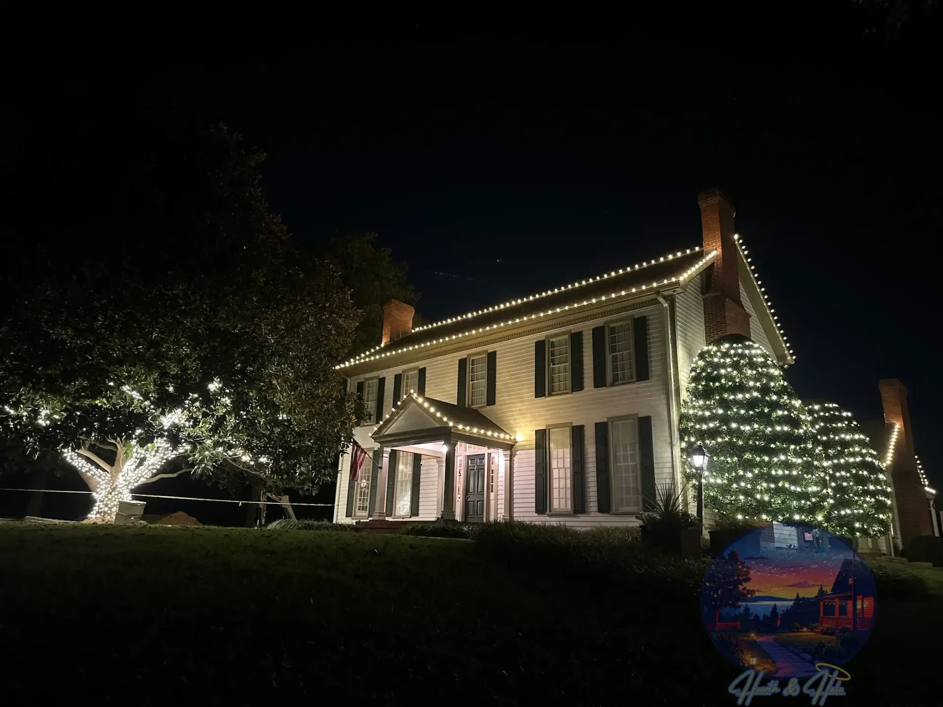 A two-story house at night illuminated with warm white string lights, accompanied by a large, light-adorned tree.